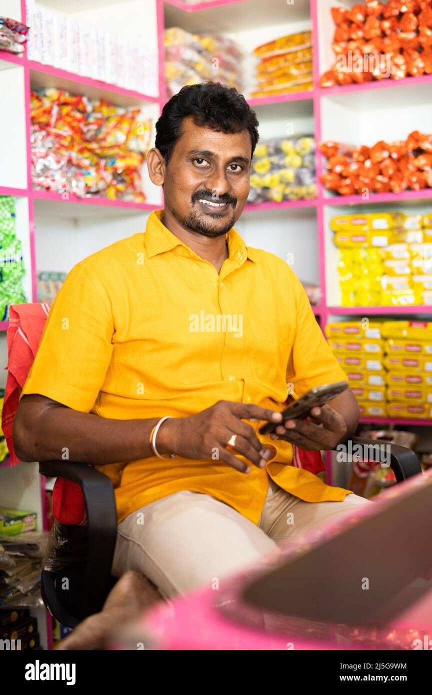 Smiling Indian shopkeeper in a yellow shirt using a smartphone in a colourful grocery store